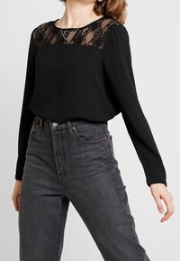 Maglietta nera a maniche lunghe con un pannello di pizzo sullo scollo. Tessuto morbido, vestibilità rilassata, abbinata a jeans grigio scuro a vita alta.