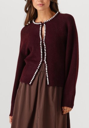 Vrouw in een donker bordeauxrode cardigan met parelknoopafwerking over een hooggemeten bruine rok, staande met één hand zichtbaar.