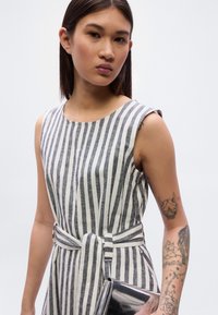 Γκρι και λευκό ριγέ αμάνικο φόρεμα με ζώνη στη μέση, κατασκευασμένο από υφή υφάσματος. Το μοντέλο κρατάει ένα γυαλιστερό μαύρο τσαντάκι χειρός.