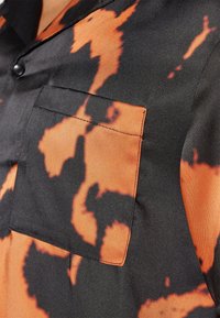Kurzarmhemd mit satinierter Oberfläche, schwarzem Grund und orangefarbenen Spritzmustern, sowie einer Brusttasche mit kontrastierenden Nähten.
