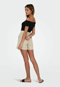 Haut noir à épaules nues, short beige à taille élastique avec poches, sandales à glissière noires plates, teint de peau lisse, debout en profil contre un fond clair.