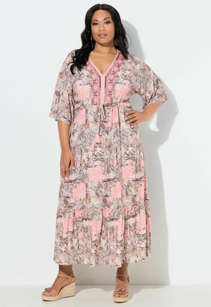 Kobieta w różowo-beżowej sukni maxi z wzorem, z rękawami do łokci i sandałami na koturnie, stojąca na białym tle.