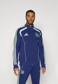 Marineblå Adidas træningsjakke med lyseblå detaljer, fuld lynlås, høj krave og tre striber på ærmerne, med et logo på brystet.