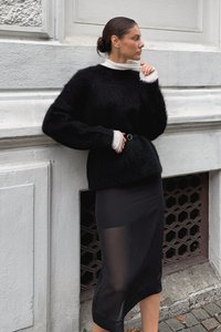 Black fuzzy sweater with a high neckline and white cuffs, paired with a black sheer skirt. Woman poses against a textured wall.