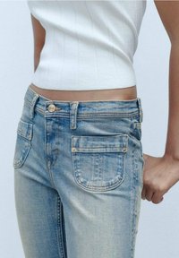 Vaqueros de mezclilla azul claro con un corte de talle medio, cinco bolsillos y una textura sutilmente desgastada. El cierre de cremallera está acompañado por un botón dorado.