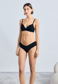 Sutien negru cu un decolteu adânc și bretele ajustabile, asortat cu chilot negru cu margini ondulate.
