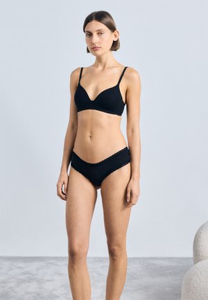 Czarna bielizna w stylu push-up z głębokim dekoltem i regulowanymi ramiączkami, w zestawie z dopasowanym czarnym majtkami z falbaniastymi brzegami.
