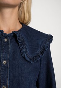Camicia in denim blu navy con colletto arricciato, chiusura con bottoni e dettagli di cucitura sottili. Il tessuto ha una finitura liscia e strutturata.