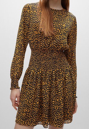 Femme portant une robe noire à manches longues avec des taches orange irrégulières et une taille et des poignets smockés, debout devant un fond uni.
