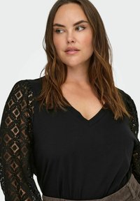 Czarny top z dekoltem w kształcie litery V i długimi rękawami wykonanymi z teksturowanej koronki z geometrycznym wzorem. Gładka tkanina stanowi kontrast dla koronki.