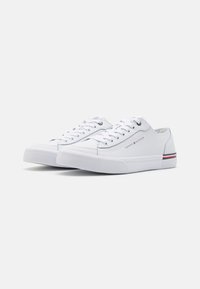 Weiße Leinensneaker mit niedrigem Profil, Schnürdesign und strukturierten Gummisohlen. Mit Logodetail und roten/blauen Streifenakzenten.