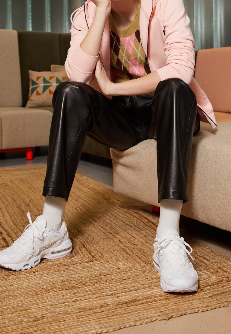 Vita sportskor, svarta fusk läderbyxor och en rosa zip-up hoodie över en mönstrad tröja. Sitter på en beige soffa med en texturerad matta.