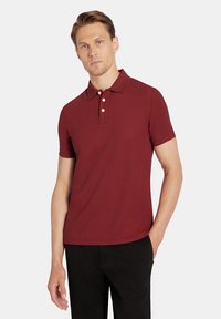 Bordowa koszulka polo z krótkim rękawem, z kołnierzykiem, trzema guzikami oraz subtelną teksturą. Zestawiona z czarnymi spodniami.