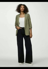 Olivovo zelený blejzer cez biely top s tenkým ramienkom, skombinovaný s čiernymi nohavicami s vysokým pásom a širokými nohavicami, doplnený čiernymi teniskami. Hladké, pohodlné textúry látok.