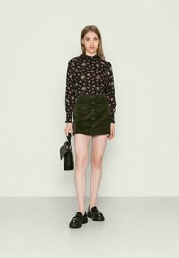 Blusa preta de flores com mangas longas, saia mini de corduroy verde com detalhes em botão, sapatos platform pretos e uma pequena mala preta.