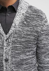 Svart och vit stickad cardigan med ett texturerat mönster, V-ringad design och två knappar framtill, med långa ärmar.