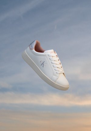 Baskets basses blanches avec doublure rose et logo Calvin Klein sur le côté, lacées, montrées sur un fond de ciel nuageux.