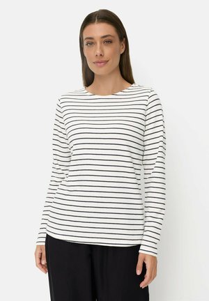 Vrouw draagt een wit shirt met lange mouwen met dunne horizontale zwarte strepen en zwarte broek tegen een effen lichte achtergrond.