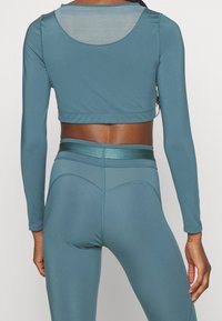 Teal long-sleeve crop top with a round back cut-out, paired with matching high-waisted leggings featuring subtle stitching details.