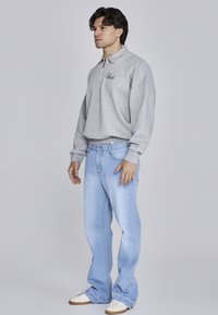 Pullover in felpa grigia chiara con collo a zip e logo ricamato; abbinato a jeans a gamba larga blu chiaro e sneakers bianche.