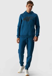 Sudadera con capucha azul y gran logotipo negro, combinada con joggers azules a juego. Material de mezcla de algodón, puños acanalados y bolsillos laterales.