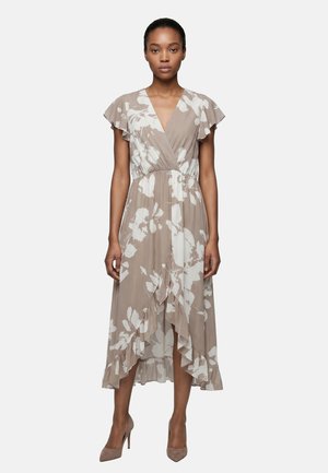 Donna che indossa un abito avvolgente beige e bianco a fiori con maniche e orlo arricciati, abbinato a scarpe beige con tacchi alti, in piedi davanti a uno sfondo semplice.