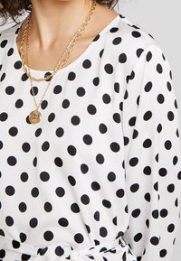 Weißes Blusenshirt mit schwarzen Punkten, mit einem runden Ausschnitt, weichem Stoff und einer gebundenen Detail an der Taille. Goldene Kettenhalskette drapiert.