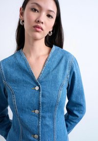 Jeune femme aux longs cheveux noirs portant des boucles d'oreilles créoles argentées et une veste en jean bleue ajustée à trois boutons sur fond blanc.