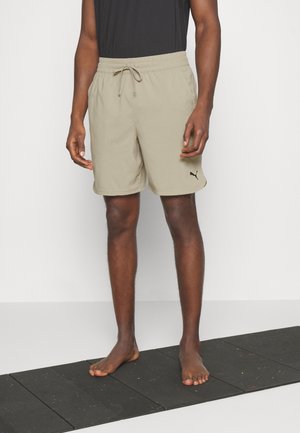 Uomo con pantaloncini beige Puma in piedi a piedi nudi su un tappetino nero da esercizio contro una parete bianca.