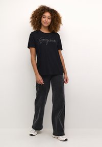 Sort bomulds t-shirt med "Gorgeous" broderet i hvid kursiv, parret med mørkegrå flared jeans og hvide sneakers. En enkel, afslappet påklædning.