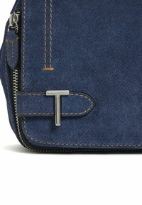 Borsa in denim blu con chiusura a zip, caratterizzata da cuciture gialle a contrasto e un accento a forma di T in tonalità argento sul davanti.