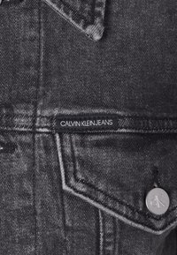 Close-up van de zak van een zwarte spijkerjasje met een "Calvin Klein Jeans" label en een metalen knoop met het gegraveerde "cK" logo.