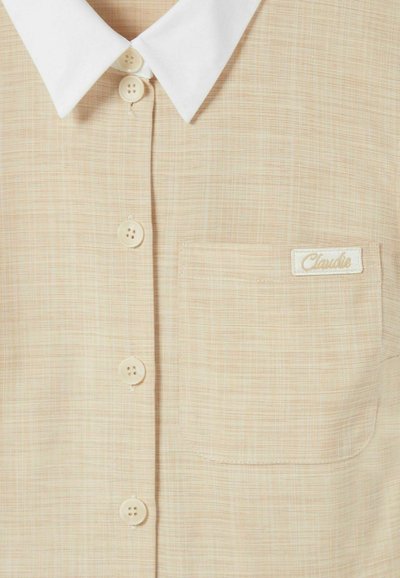 Chemise beige à boutons avec un col blanc, en tissu texturé, dotée de quatre boutons clairs et d'un petit patch logo sur la poche gauche.