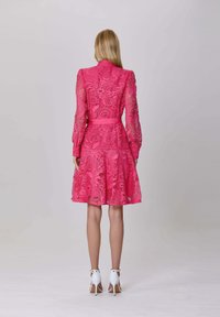 Femme de dos, portant une robe en dentelle rose à manches longues avec une ceinture et des chaussures à talons hauts blanches, sur un fond uni.