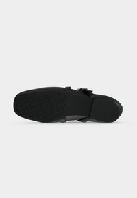 Chaussure noire avec une semelle en caoutchouc texturée comportant un motif géométrique et une petite bride à boucle sur le dessus.