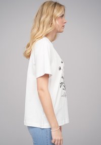 T-shirt en coton blanc à manches courtes, présentant un imprimé graphique floral noir et du texte. Design simple avec une coupe décontractée. Jean en denim bleu clair montré.