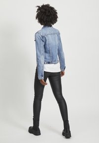 Světle modrá džínová bunda s přiléhavým střihem, vybavená límcem, dlouhými rukávy a dvěma předními kapsami s klopami. Natažená přes bílou košili a černé legíny.