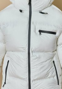 Doudoune blanche avec capuche, dotée d'une fermeture éclair grise et d'accents noirs. Comprend des poches zippées sur la poitrine et sur les côtés.