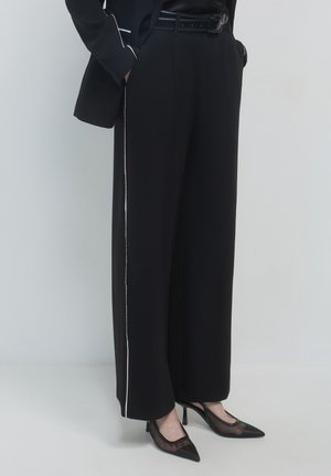 Woman wearing black wide-leg trousers with white side stripes, black belt, black blazer with white piping, and black sheer pointed heels.