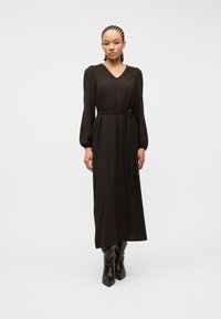 Γυναίκα με πλεξούδες, φορεμένη με μαύρο μίντι φόρεμα με μακριά μανίκια και ζώνη στη μέση, καθώς και με σκουρόχρωμες μπότες μυτερής μύτης, στέκεται μπροστά σε μια απλή φόντο.