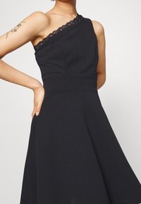 Černé šaty s designem na jedno rameno, krajkovým lemem, vypasovaným živůtkem a rozšířenou sukní s ukrytými kapsami. Hladká textura látky.