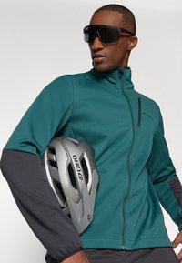 Türkise Zip-Jacke mit dunkelgrauen Ärmeln, leichtem Stoff und schlanker Passform. Schwarze Sonnenbrille und ein grauer Giro-Helm unter dem Arm gehalten.