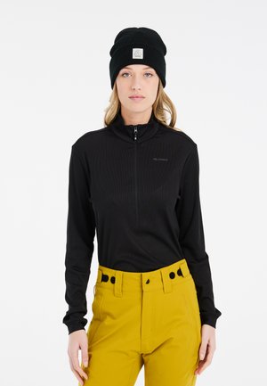 Zwarte rits-tot aan de hals lange mouwen top met ribbelstructuur, gecombineerd met gele high-waisted broek met zwarte knoopaccenten en een bijpassende beanie.