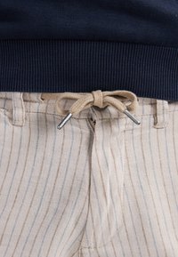 Pantaloni beige a strisce con strisce blu e marroni, dotati di un laccio beige annodato e punte in argento. Maglione a coste blu navy sopra.
