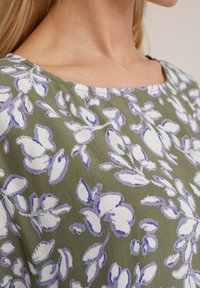Blusa floreale caratterizzata da un tessuto verde con fiori bianchi e lavanda, scollatura rotonda e una superficie morbida e testurizzata.