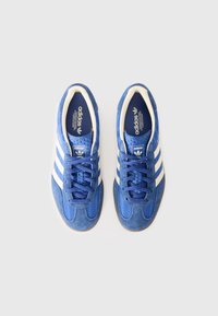 Încălțăminte sport bleu din piele de căprioară cu dungi albe, partea superioară texturată și design cu șireturi. Include un guler căptușit și o talpă din cauciuc de tip gum.