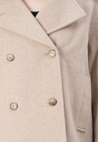 Beige villa kaksoisnapitakki, jossa on neljä nappia ja leveät kaulukset, sivutasku ja musta vaatetus alla.