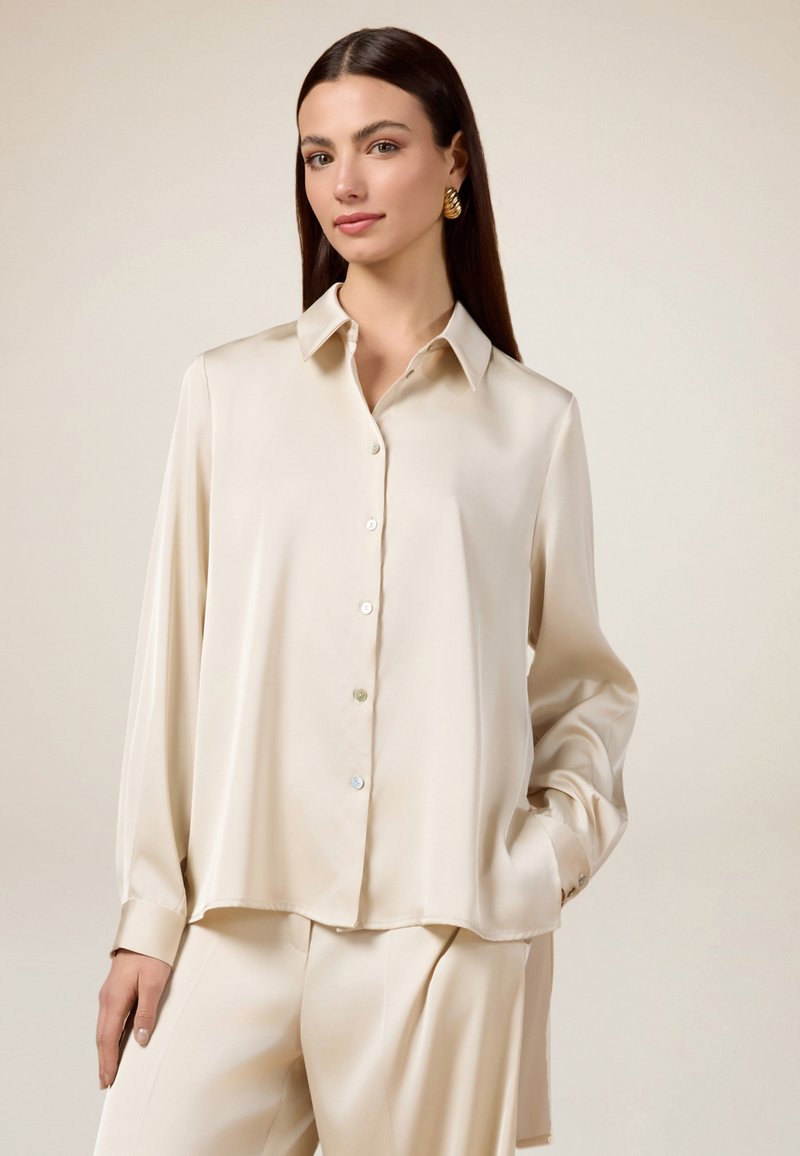 Camicia lunga in satin crema con maniche lunghe, colletto classico, chiusura con bottoni frontale e spacchi laterali, caratterizzata da un tessuto lucido e una vestibilità rilassata.