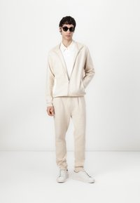 Veste beige zippée avec un motif subtil, polo blanc en dessous, pantalon assorti et baskets blanches. Modèle portant des lunettes de soleil noires.