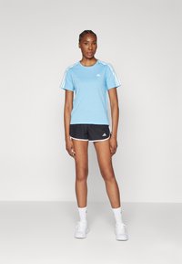 Svijetloplava sportska majica s bijelim trostrukim detaljima na ramenima, u kombinaciji s crnim kratkim hlačama koje imaju bijele akcentne i Adidas logo.